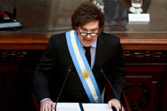 Político argentino faz discurso enérgico, usando faixa presidencial azul e branca, em ambiente governamental com microfones.