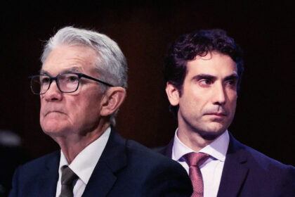 Dois homens de terno, um mais velho usando óculos, outro mais novo, aparecem em um contexto sério ou formal.