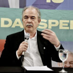 Homem de terno preto em coletiva de imprensa, falando ao microfone, com fundo de banner colorido e copo de água na mesa.