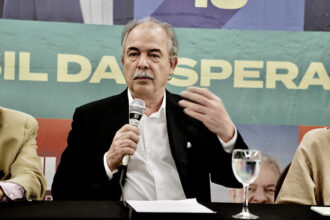 Homem de terno preto em coletiva de imprensa, falando ao microfone, com fundo de banner colorido e copo de água na mesa.