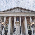 Fachada do Royal Exchange em Londres com colunas neoclássicas, esculturas, lâmpadas e céu nublado ao fundo.
