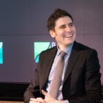 Homem em terno sorrindo durante evento corporativo, em frente a telas com gráfico de crescimento ao fundo.