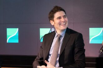 Homem em terno sorrindo durante evento corporativo, em frente a telas com gráfico de crescimento ao fundo.
