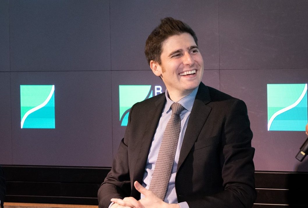 Homem em terno sorrindo durante evento corporativo, em frente a telas com gráfico de crescimento ao fundo.