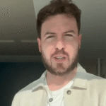 Pessoa de barba se encontra em ambiente interno iluminado pelo sol, usando camisa clara, ao lado de uma parede azul.