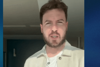 Pessoa de barba se encontra em ambiente interno iluminado pelo sol, usando camisa clara, ao lado de uma parede azul.