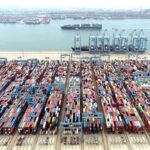 Porto movimentado com dezenas de contêineres coloridos organizados em filas, guindastes e navios ao fundo no cais.