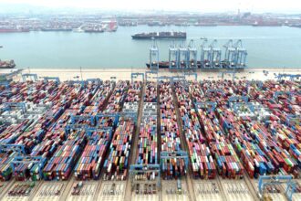 Porto movimentado com dezenas de contêineres coloridos organizados em filas, guindastes e navios ao fundo no cais.