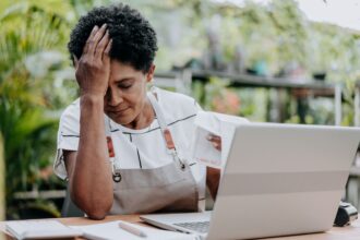 Pessoa em um ambiente de trabalho, aparentando estresse, com a mão na cabeça, ao lado de um laptop e documentos.