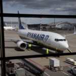 Avião estacionado no aeroporto com funcionários em uniformes refletivos próximos, bags e equipamento de solo visíveis.