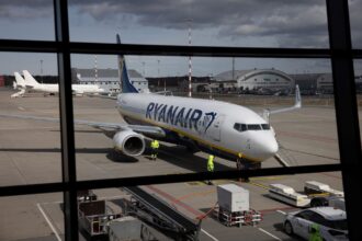 Avião estacionado no aeroporto com funcionários em uniformes refletivos próximos, bags e equipamento de solo visíveis.