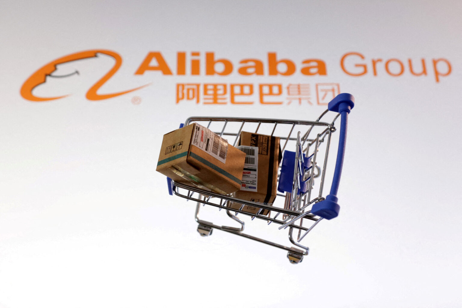 Carrinho de compras em miniatura com caixas de papelão, destacado sobre o fundo com o logotipo do Alibaba Group.