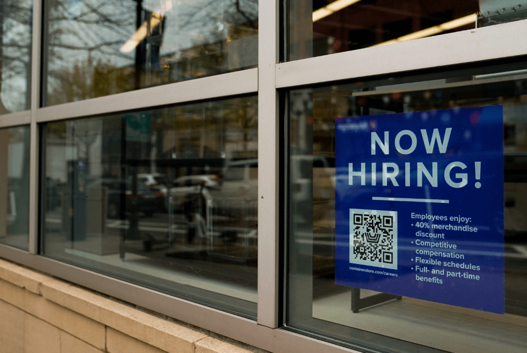 Cartaz de Now Hiring em uma vitrine, destacando benefícios como descontos em mercadorias e horários flexíveis.