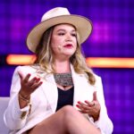 Mulher de chapéu e blazer branco gesticula durante palestra, fundo roxo com luzes, microfone auricular.