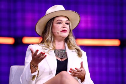 Mulher de chapéu e blazer branco gesticula durante palestra, fundo roxo com luzes, microfone auricular.