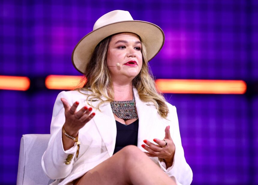 Mulher de chapéu e blazer branco gesticula durante palestra, fundo roxo com luzes, microfone auricular.