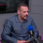 Homem de camisa jeans participa de entrevista em estúdio de rádio, falando ao microfone azul. Ambiente moderno.
