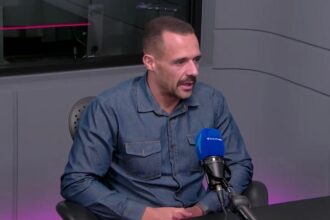 Homem de camisa jeans participa de entrevista em estúdio de rádio, falando ao microfone azul. Ambiente moderno.