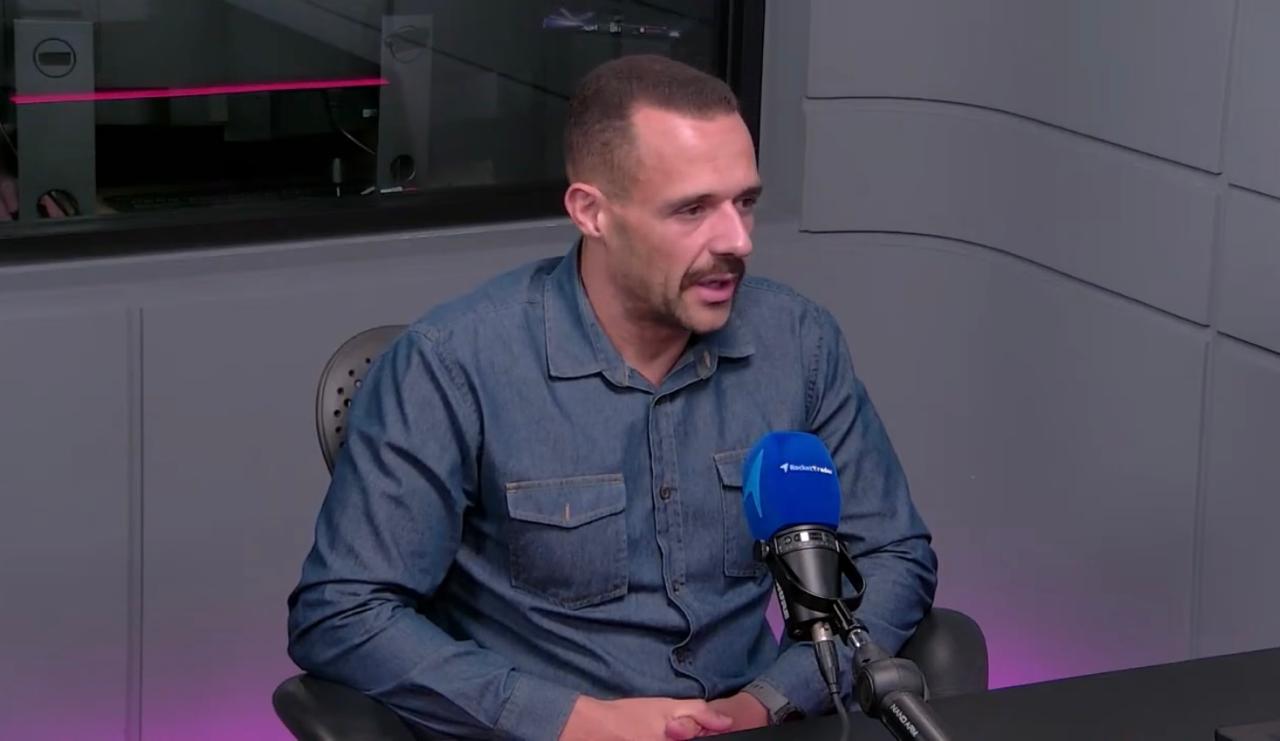 Homem de camisa jeans participa de entrevista em estúdio de rádio, falando ao microfone azul. Ambiente moderno.