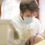 Dentista atendendo paciente em um consultório, usando máscara e luvas, realizando procedimento odontológico com instrumentos.