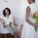 Criança presenteia mulher com flores em sala. Mulher sorri sentada em poltrona, criança esconde buquê de tulipas laranja.
