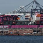 Navio cargueiro com contêineres coloridos atracado em porto, guindastes ao fundo, mostrando movimentação portuária.