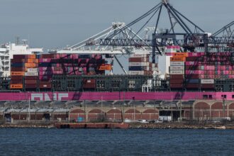 Navio cargueiro com contêineres coloridos atracado em porto, guindastes ao fundo, mostrando movimentação portuária.