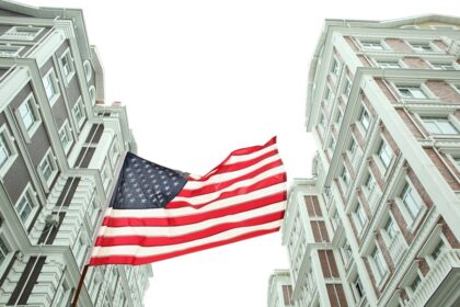 Bandeira dos EUA tremulando entre edifícios altos em um ambiente urbano, destacando arquitetura clássica e patriotismo.