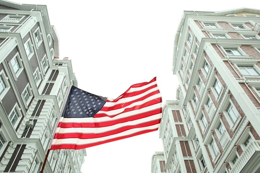 Bandeira dos EUA tremulando entre edifícios altos em um ambiente urbano, destacando arquitetura clássica e patriotismo.