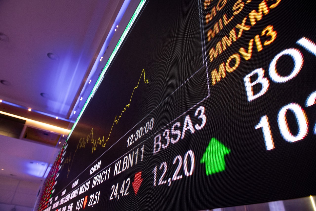 Painel digital de cotações da bolsa de valores com gráficos de ações e indicadores econômicos em destaque.