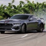 Carro esportivo cinza fazendo drift em pista sinuosa, com fumaça saindo dos pneus. Vegetação verde ao fundo.