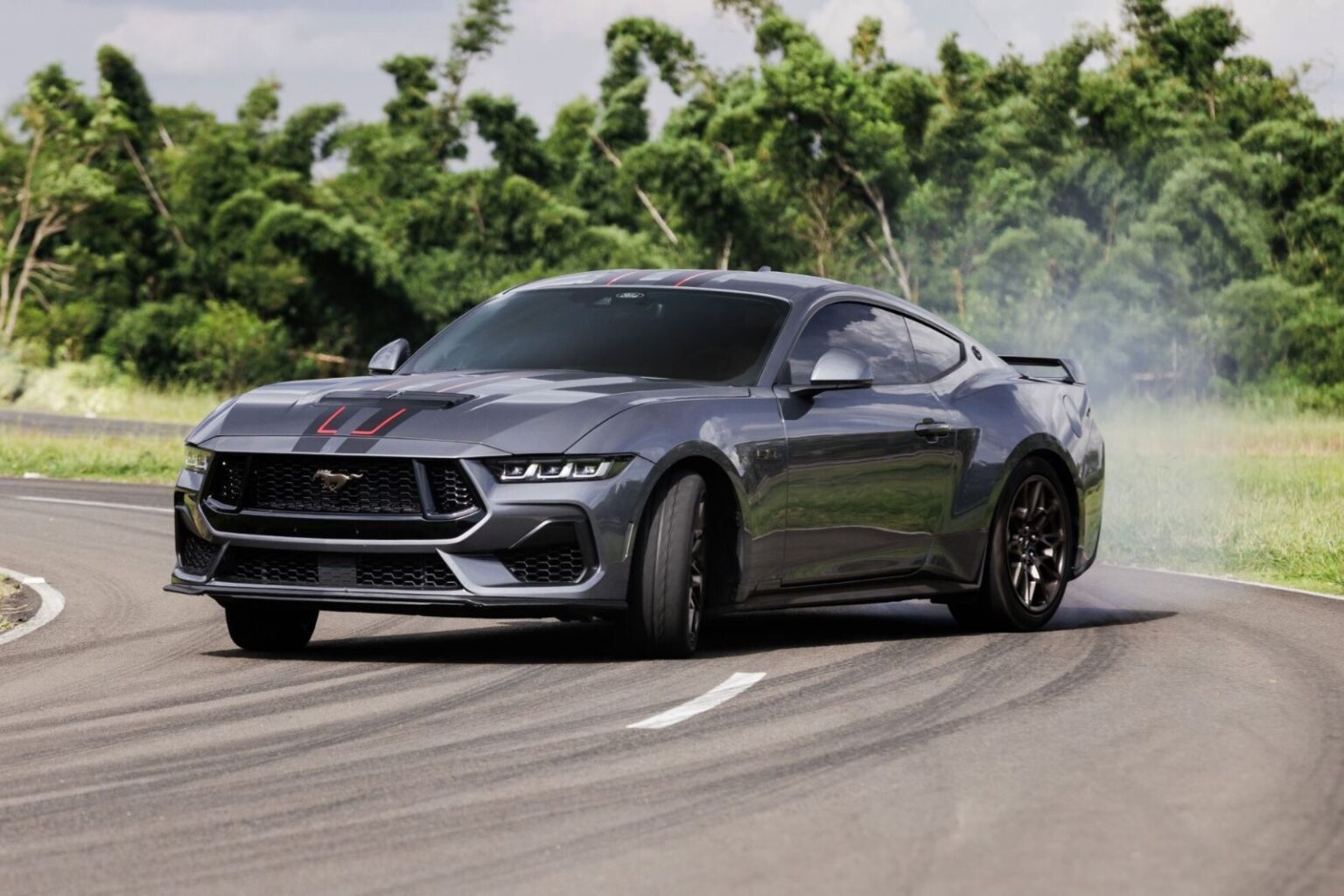 Carro esportivo cinza fazendo drift em pista sinuosa, com fumaça saindo dos pneus. Vegetação verde ao fundo.