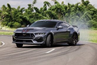 Carro esportivo cinza fazendo drift em pista sinuosa, com fumaça saindo dos pneus. Vegetação verde ao fundo.