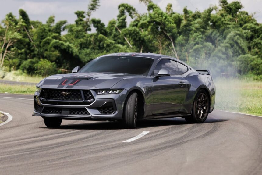 Carro esportivo cinza fazendo drift em pista sinuosa, com fumaça saindo dos pneus. Vegetação verde ao fundo.