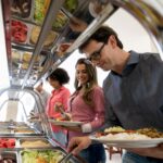 Pessoas servindo-se em buffet de comida variada em restaurante, com pratos coloridos e opções de saladas frescas.