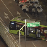 Ônibus verde elétrico em rodovia à noite, cercado por policiais e viaturas, próximo a uma placa de retorno para Itaquera.