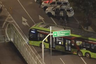 Ônibus verde elétrico em rodovia à noite, cercado por policiais e viaturas, próximo a uma placa de retorno para Itaquera.