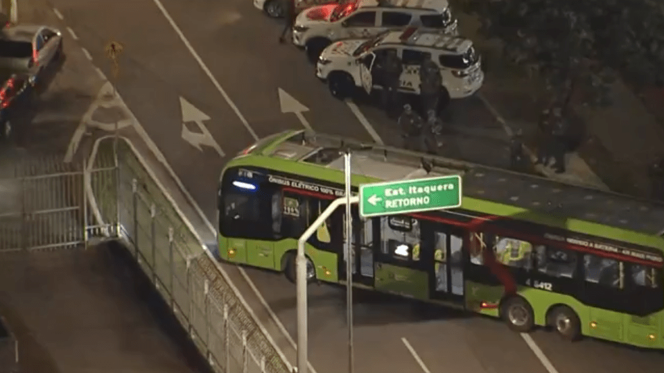 Ônibus verde elétrico em rodovia à noite, cercado por policiais e viaturas, próximo a uma placa de retorno para Itaquera.
