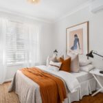 Quarto moderno com cama de solteiro, colcha laranja, almofadas e abajur em um ambiente iluminado por luz natural.