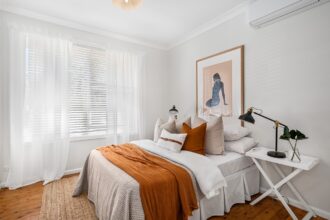 Quarto moderno com cama de solteiro, colcha laranja, almofadas e abajur em um ambiente iluminado por luz natural.