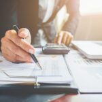 Profissional analisa gráficos e números, usando calculadora e laptop. Ambiente de escritório, foco em planejamento financeiro.