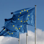 Bandeiras da União Europeia tremulando ao vento, destacando doze estrelas douradas em um céu azul, simbolizando unidade.