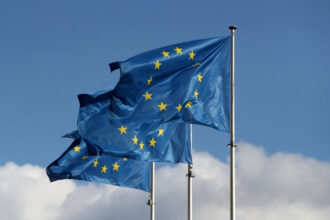 Bandeiras da União Europeia tremulando ao vento, destacando doze estrelas douradas em um céu azul, simbolizando unidade.