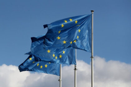 Bandeiras da União Europeia tremulando ao vento, destacando doze estrelas douradas em um céu azul, simbolizando unidade.