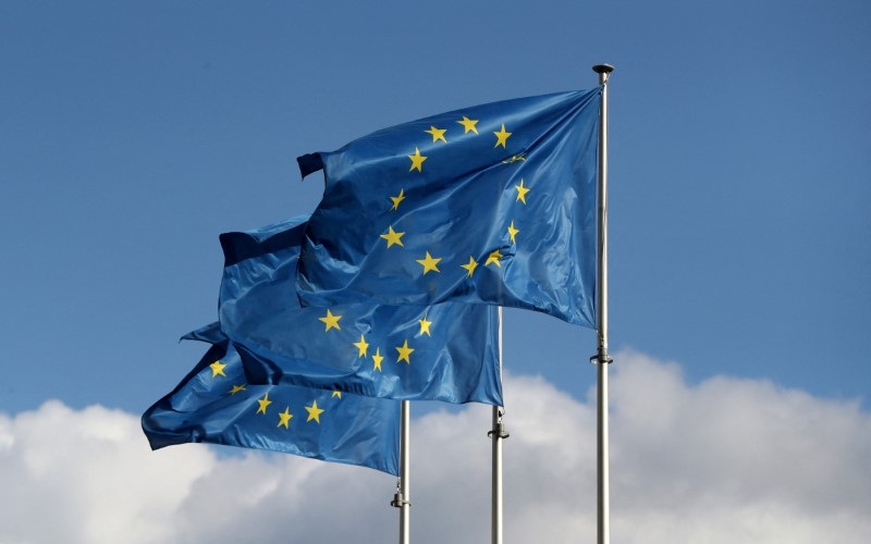 Bandeiras da União Europeia tremulando ao vento, destacando doze estrelas douradas em um céu azul, simbolizando unidade.