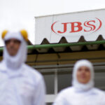 Funcionários em uniformes brancos em frente ao prédio da JBS, destaque para logotipo vermelho no topo da construção.