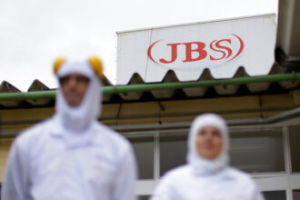 Funcionários em uniformes brancos em frente ao prédio da JBS, destaque para logotipo vermelho no topo da construção.