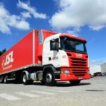 Caminhão vermelho e branco da JSL em pátio de logística sob céu azul, transportando carga com eficiência.