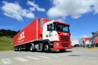 Caminhão vermelho e branco da JSL em pátio de logística sob céu azul, transportando carga com eficiência.
