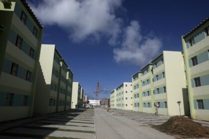 Conjunto habitacional com prédios residenciais em tons de verde, céu azul, e uma grua ao fundo.